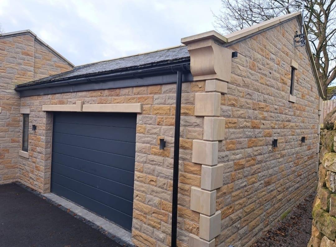 A finished detached garage in natural sandstone with dressed quoins, a stone lintel, slate roof and a dark sectional garage door.