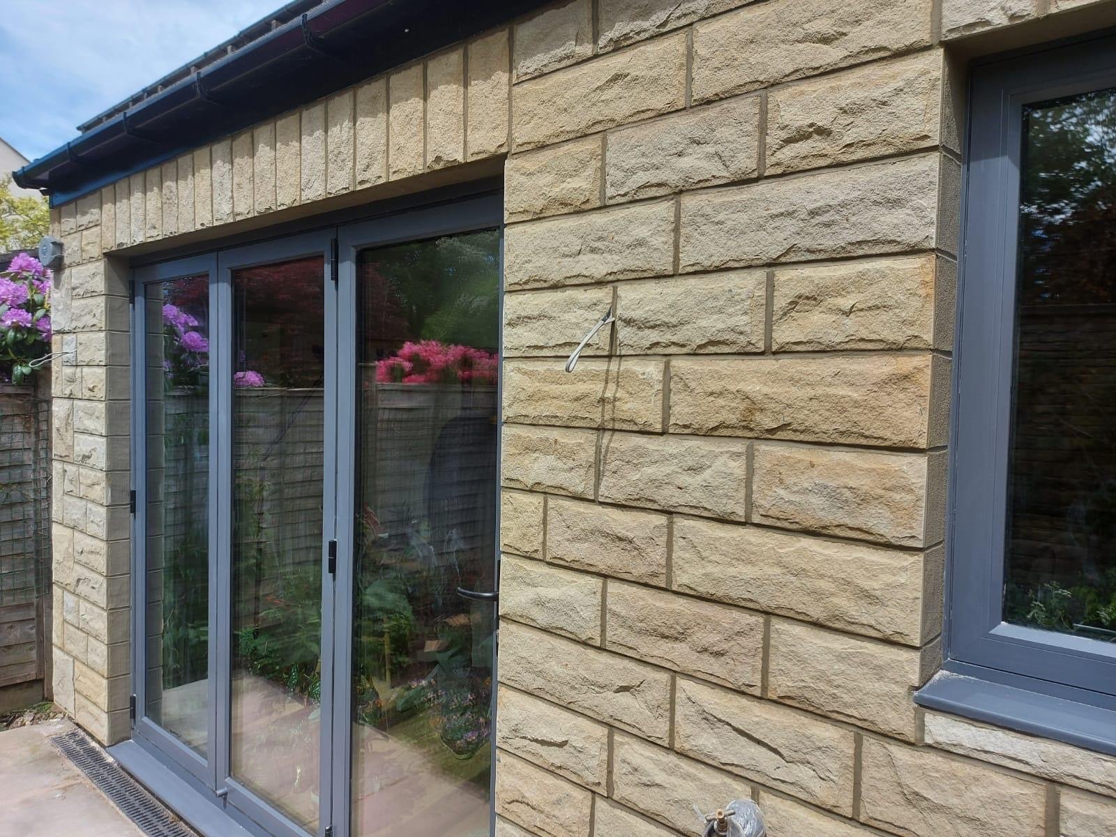 Close-up of a finished stone extension elevation in coursed sandstone with grey aluminium bi-fold doors, under a blue sky.